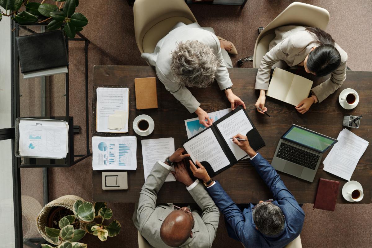 Group of middle aged multiethnic business professionals collaborating around table reviewing documents and using laptop top view showing teamwork and corporate meeting environment