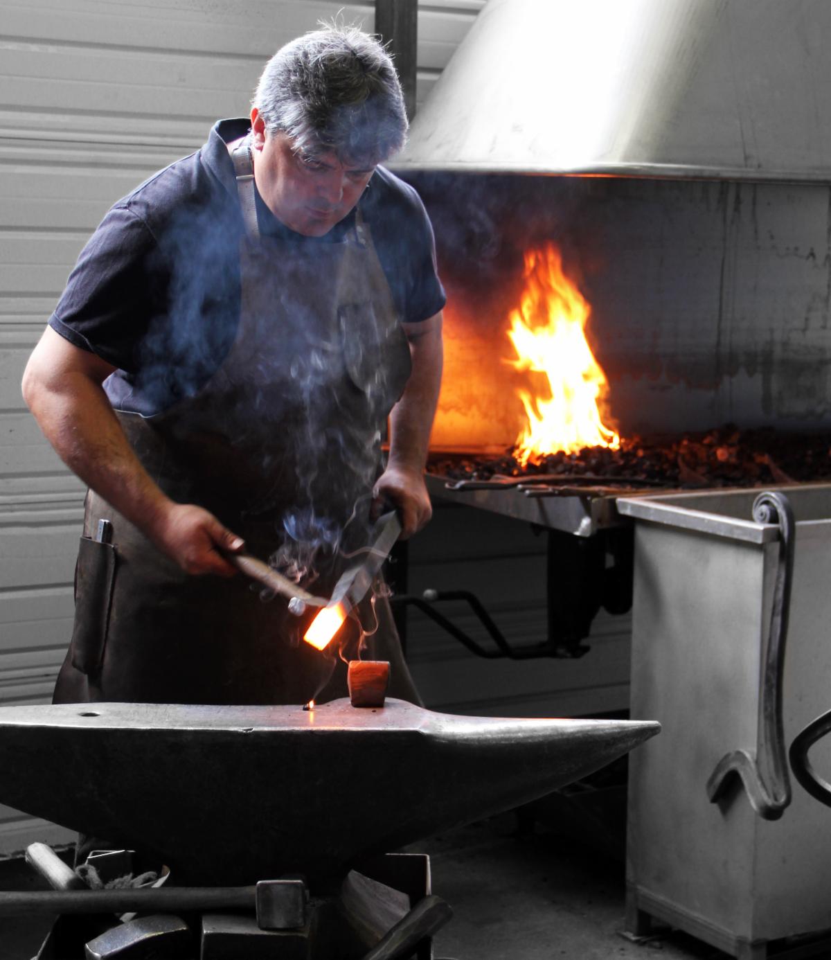 A white male with gray hair wearing a t shirt and an apron forges steel in a foundry. He uses tools to work with a piece of hot metal on a black table, with a furnace burning in behind him.