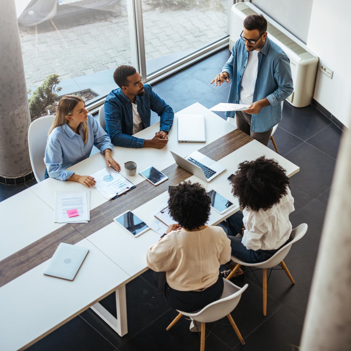 A diverse group of professionals in a modern office setting engaged in a collaborative business meeting. Team members are actively discussing strategies and projects with the aid of technology and paperwork.