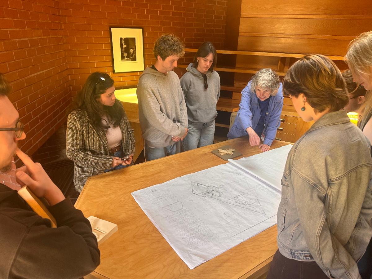 Group of volunteers and staff gathered around a large table inside the Frank Lloyd Wright House in Ebsworth Park examining architectural drawings and small wooden models during a hands on training or discussion in a warm wood paneled room.
