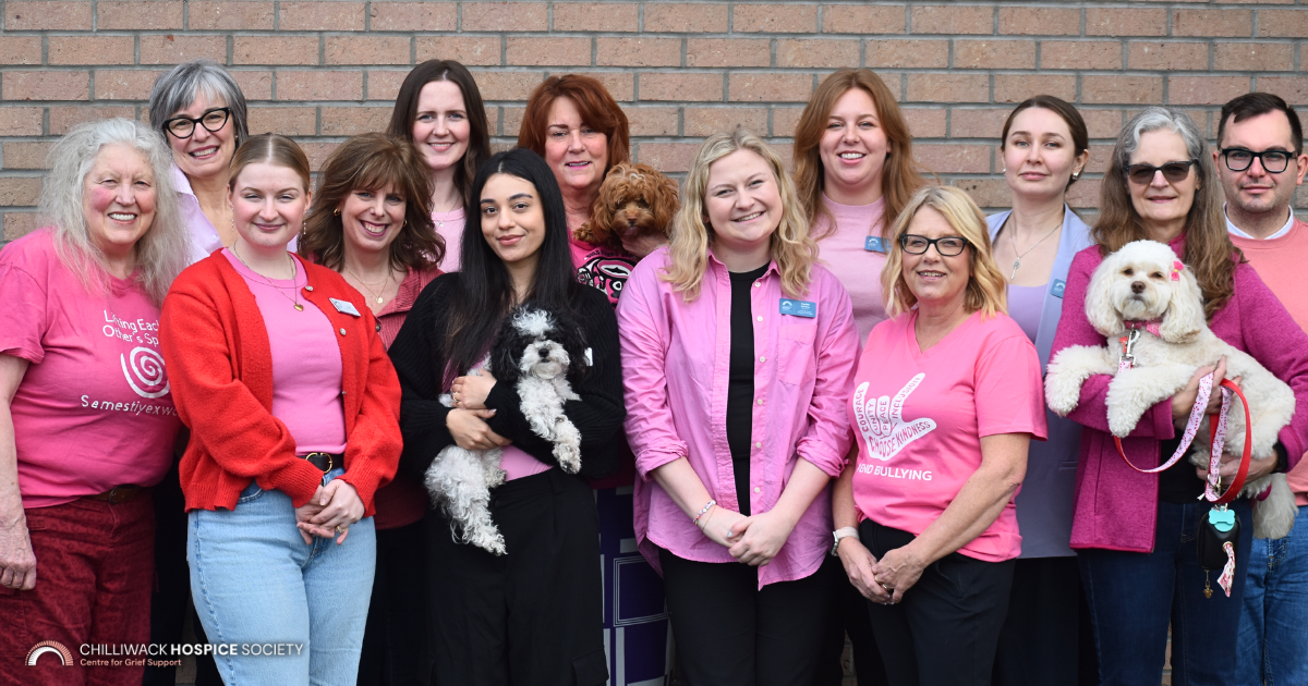 Large group of Chilliwack Hospice Society staff gathered in front of a brick wall wearing pink shirts and sweaters for Pink Shirt Day. Two small dogs are being held in the front row, and one in the back. The team stands closely together, smiling in support of anti-bullying and kindness.