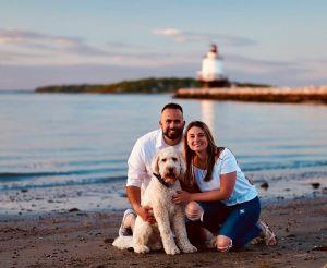 Smiling man and woman with white dog on a beach with lighthouse in background.