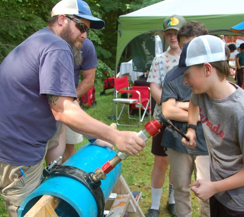 Bearded man in blue shirt helping student drill a hole in a blue pipe.