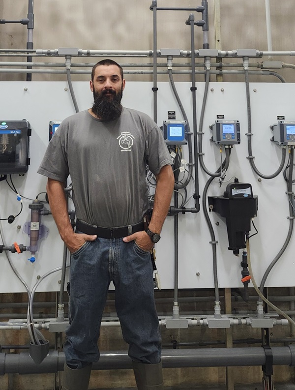 Tall man with dark brown hair and beard standing in front of a white wall lined with meters and pipes.
