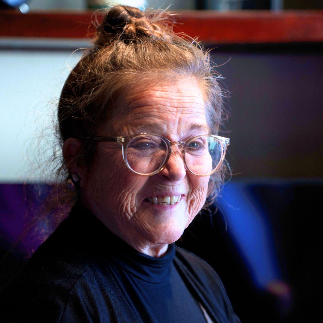 Close up of an older woman with glasses smiling with her hair in a bun and wearing a dark top