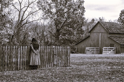 Black and white image of an old barn and woman standing with her hands behind her back in pioneer dress