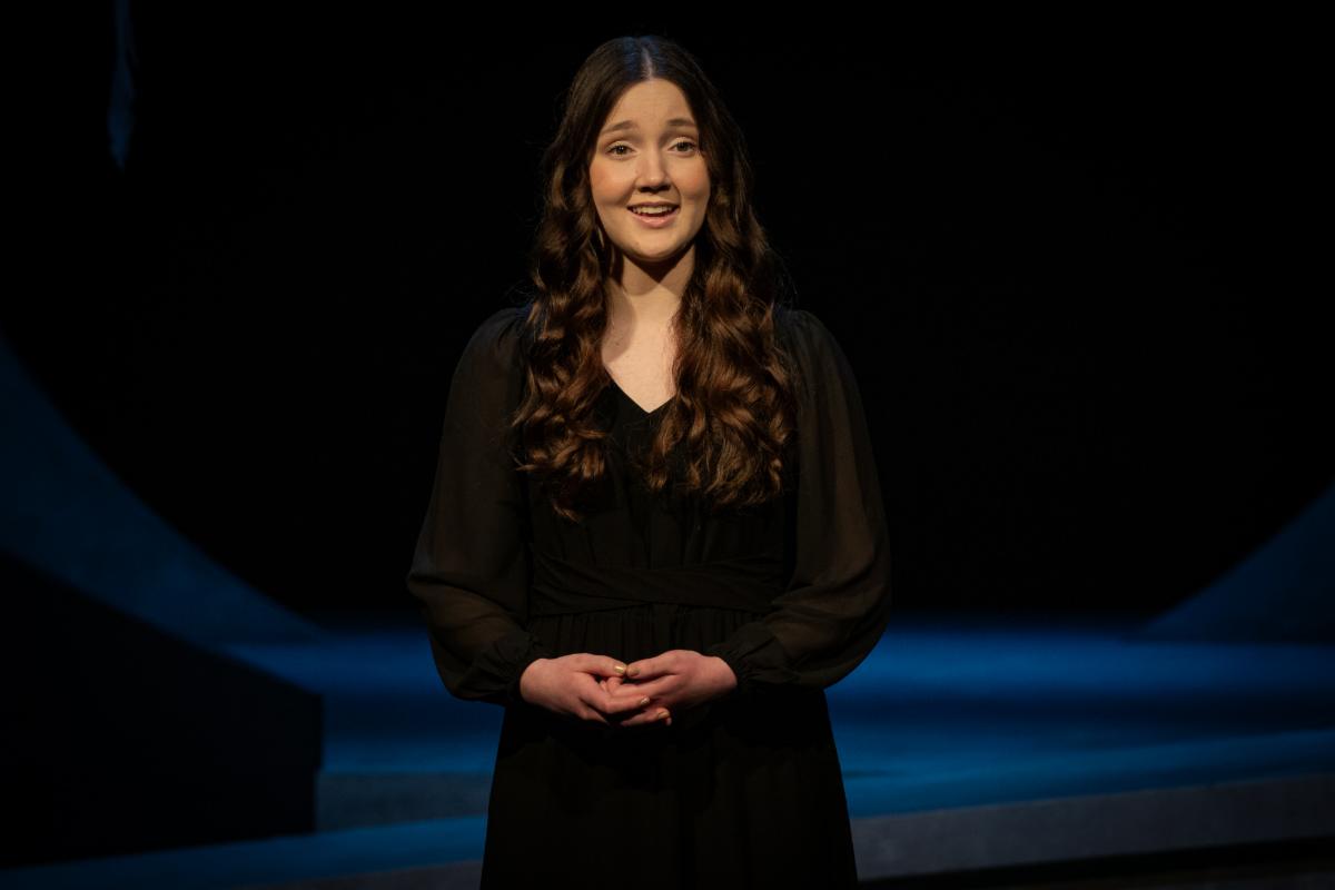 Sydney Frost was named the 2025 Washington State Poetry Out Loud Champion. She is pictured on stage in a black dress with her hands folded in front of her as she recites a poem.