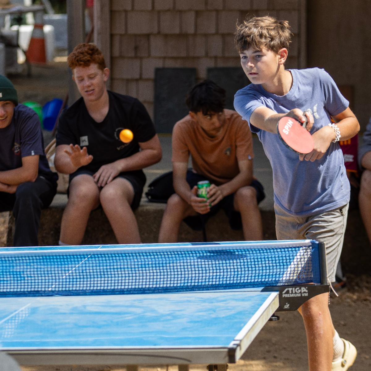 a youth in motion playing ping pong with kids in the background watching. 