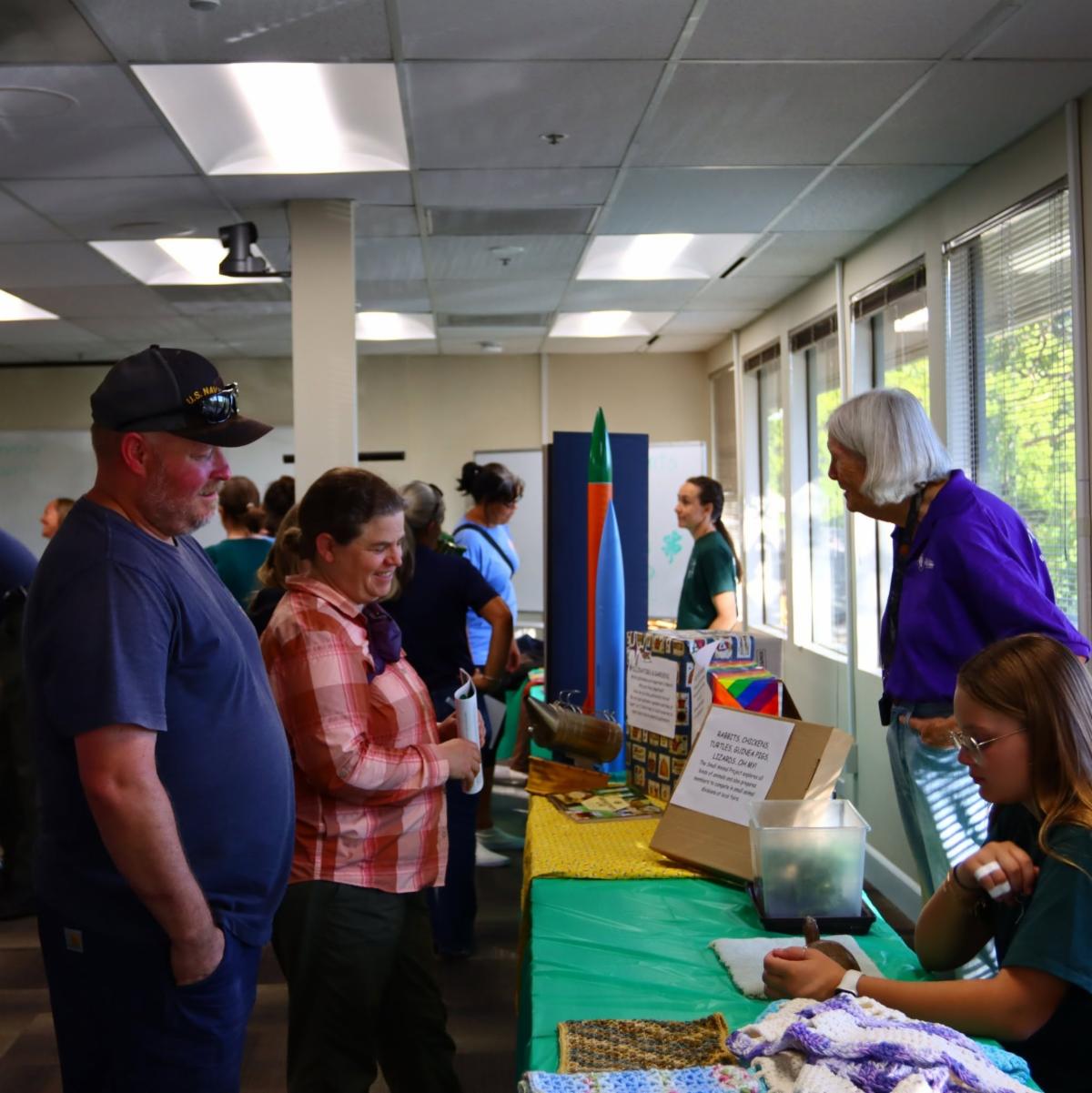 people on either side of a table with different materials such as rockets, a pet turtle, and poster boards displayed on the table. Showcase event. 
