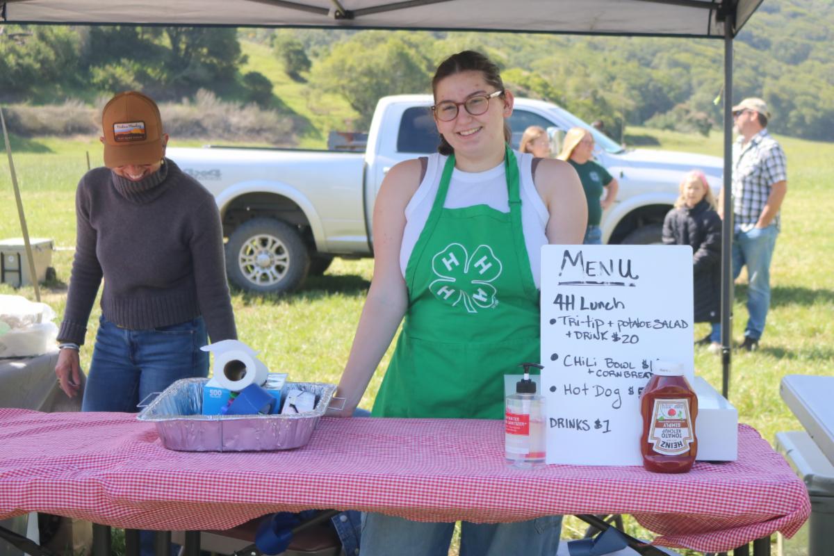 a woman in a 4-H green apron at a BBQ stand smiling 