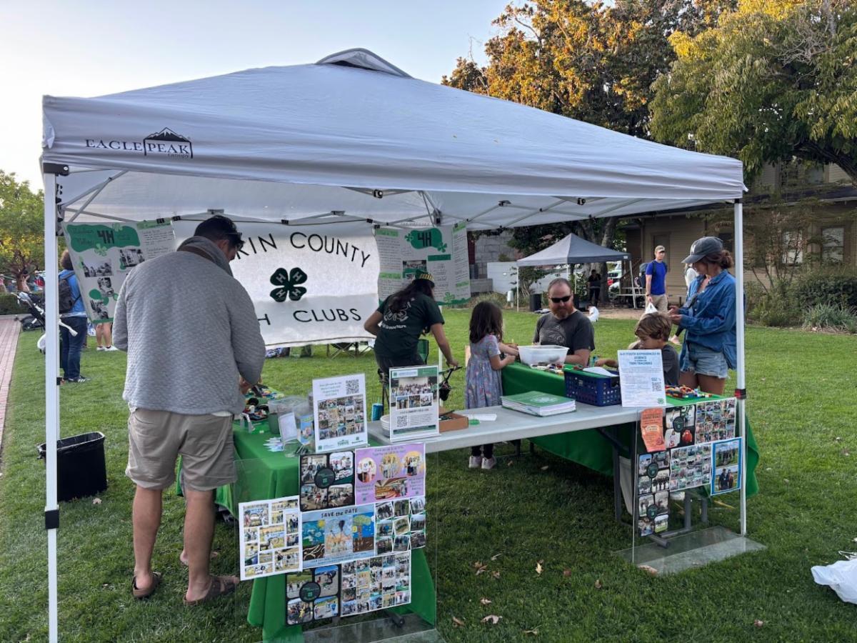 a white canopy set up with tables and materials to promote 4-H material and the 4-H program. A white banner with green lettering that says "Marin County 4-H Clubs" is hanging in the foreground with many people surrounding the booth