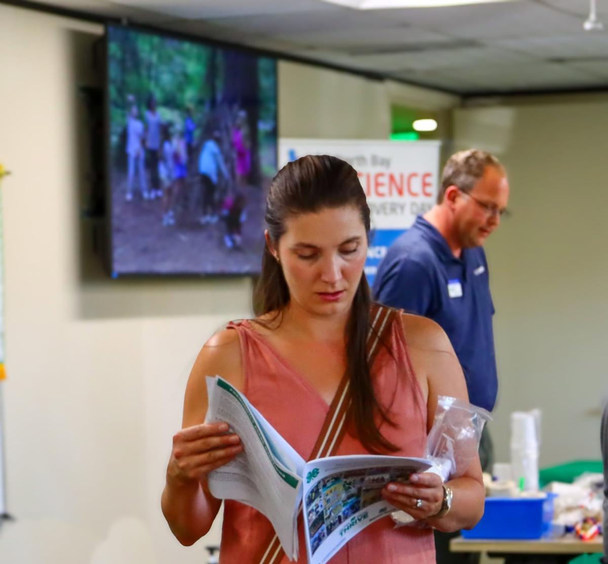 a woman reading through a booklet with a tv screen in the background at the 4-H open house event. 