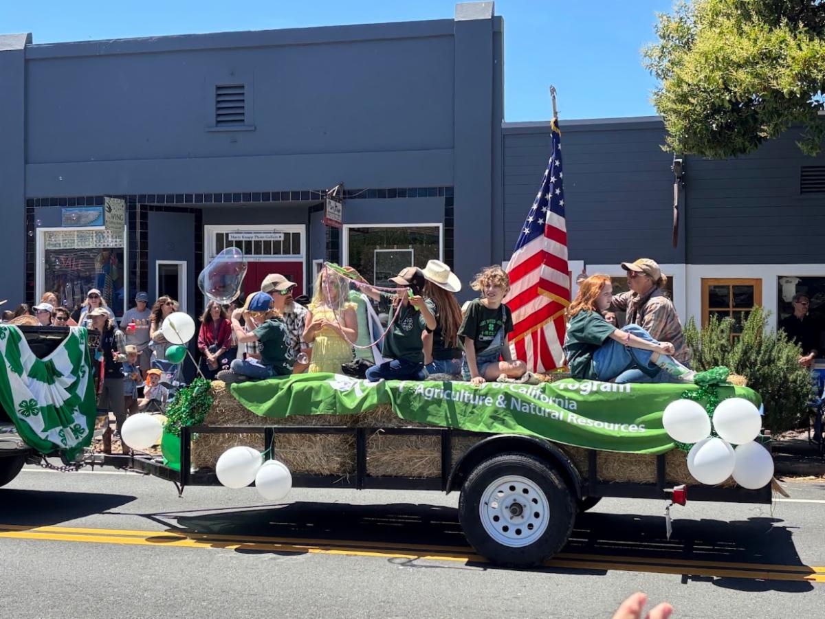 a group of kids in a 4-H parade float sitting on top of hay barrels. one of the children has a large bubble wand. The float is decorated with white and green balloons with a 4-H youth development table cloth hanging over the edge and an American flag.