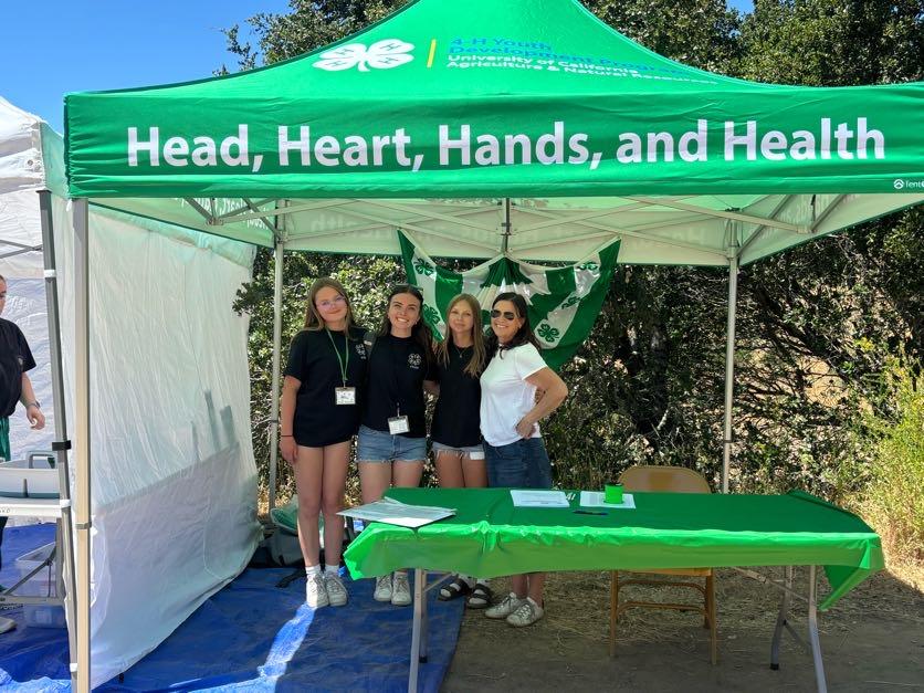 4 people underneth a green 4-H canopy tent with the words "Head, Heart, Hands and Health" with a check in table. 