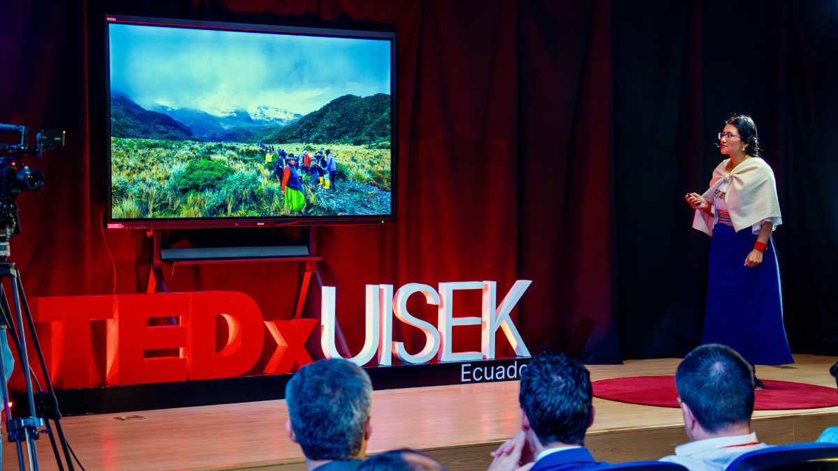 Female speaker in traditional attire presents on stage at TEDxUISEK Ecuador, standing beside a large screen displaying a scenic mountain landscape with people hiking.