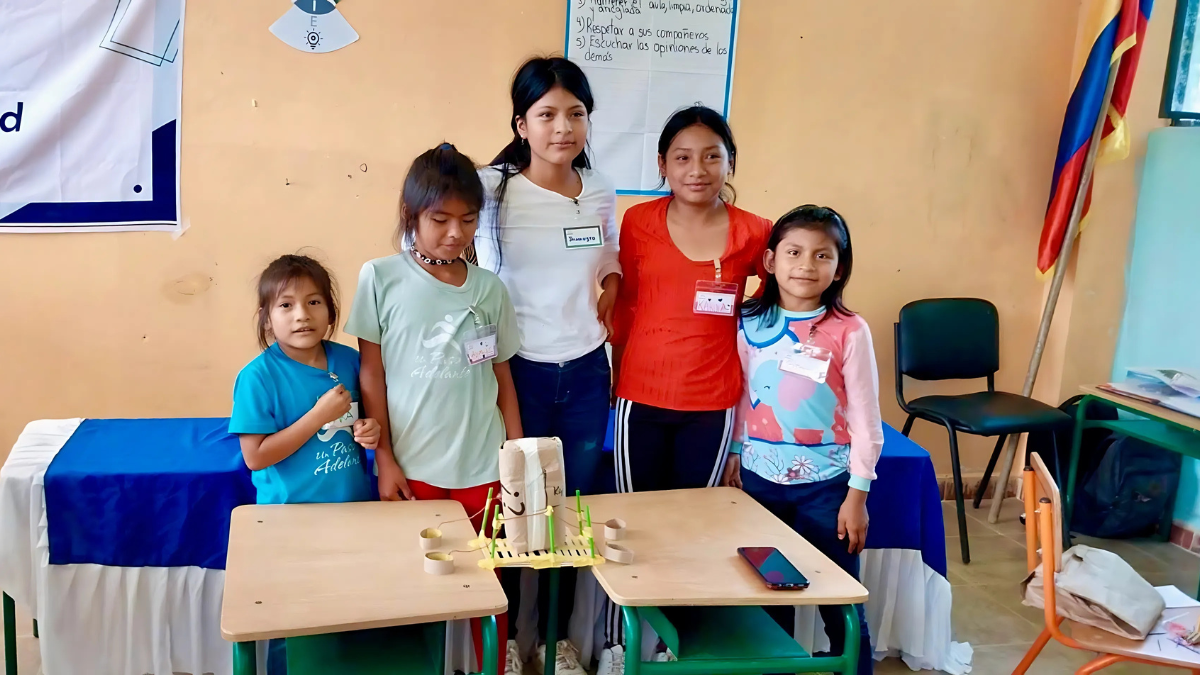 Group of five smiling girls standing behind a classroom desk displaying a handmade STEM project with paper rolls and straws, in a brightly lit educational setting decorated with posters and flags