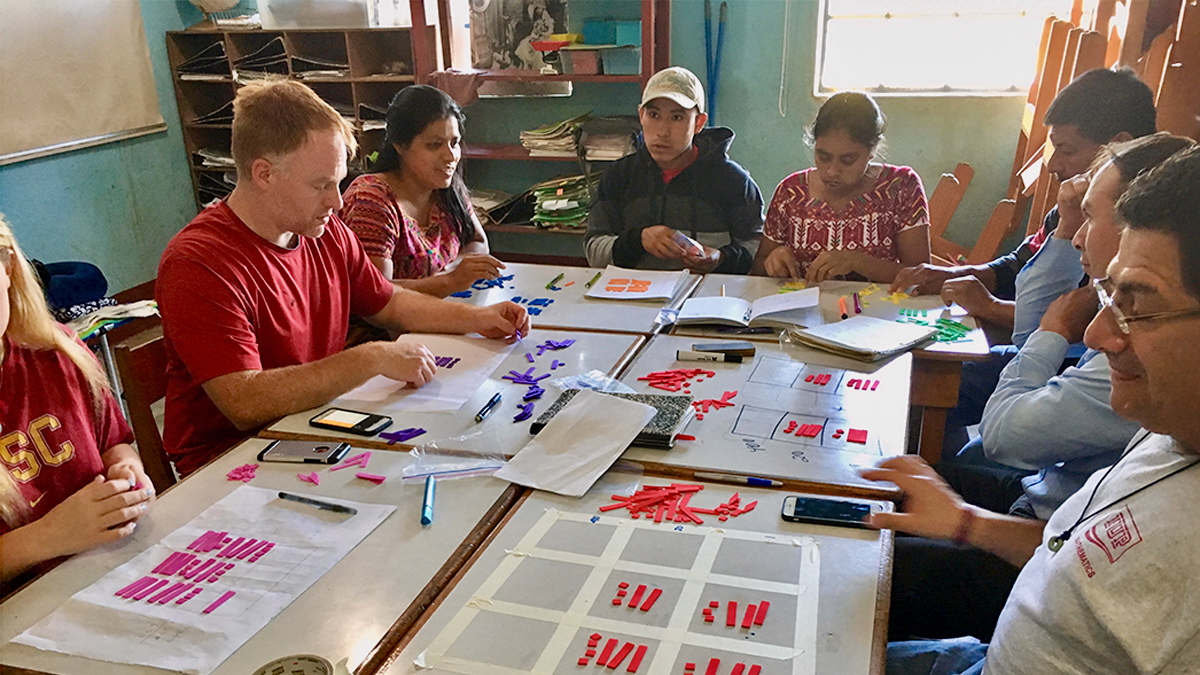 Teachers collaborate around classroom tables using colorful math manipulatives and chart papers during a hands on training session