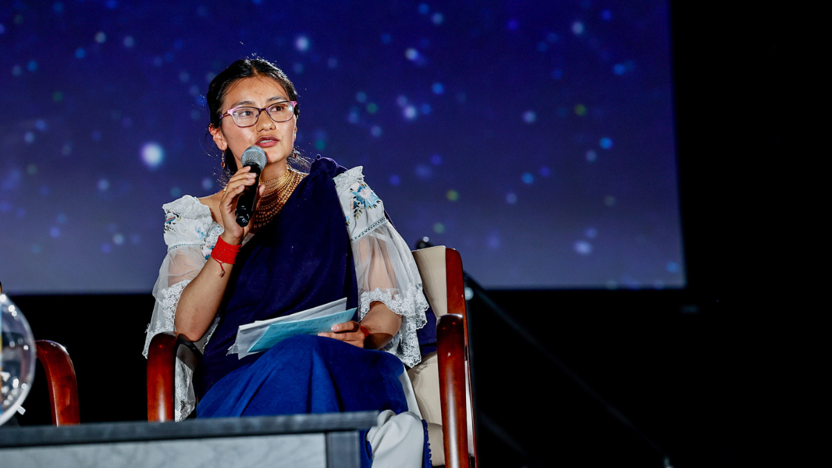Woman wearing traditional clothing and glasses speaks into a microphone while seated on stage, holding notes against a dark, starry background.