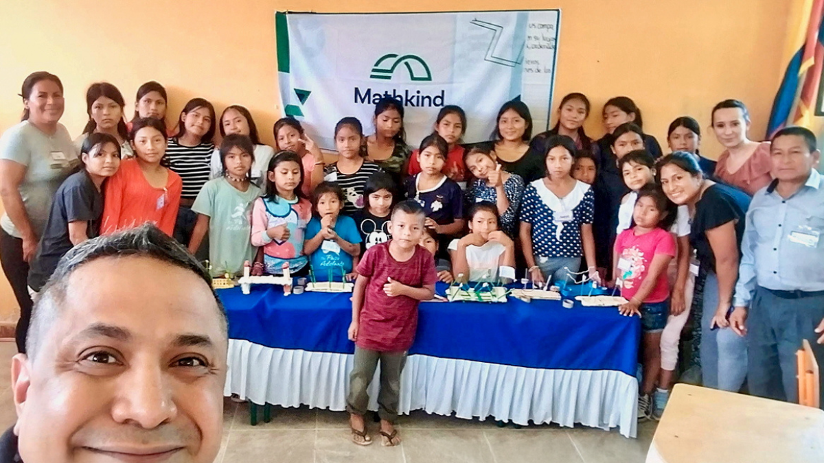 Large group of smiling children and adults posing in a classroom around a table filled with handmade STEM projects, with a “Mathkind” banner in the background and one man taking a cheerful selfie in the foreground