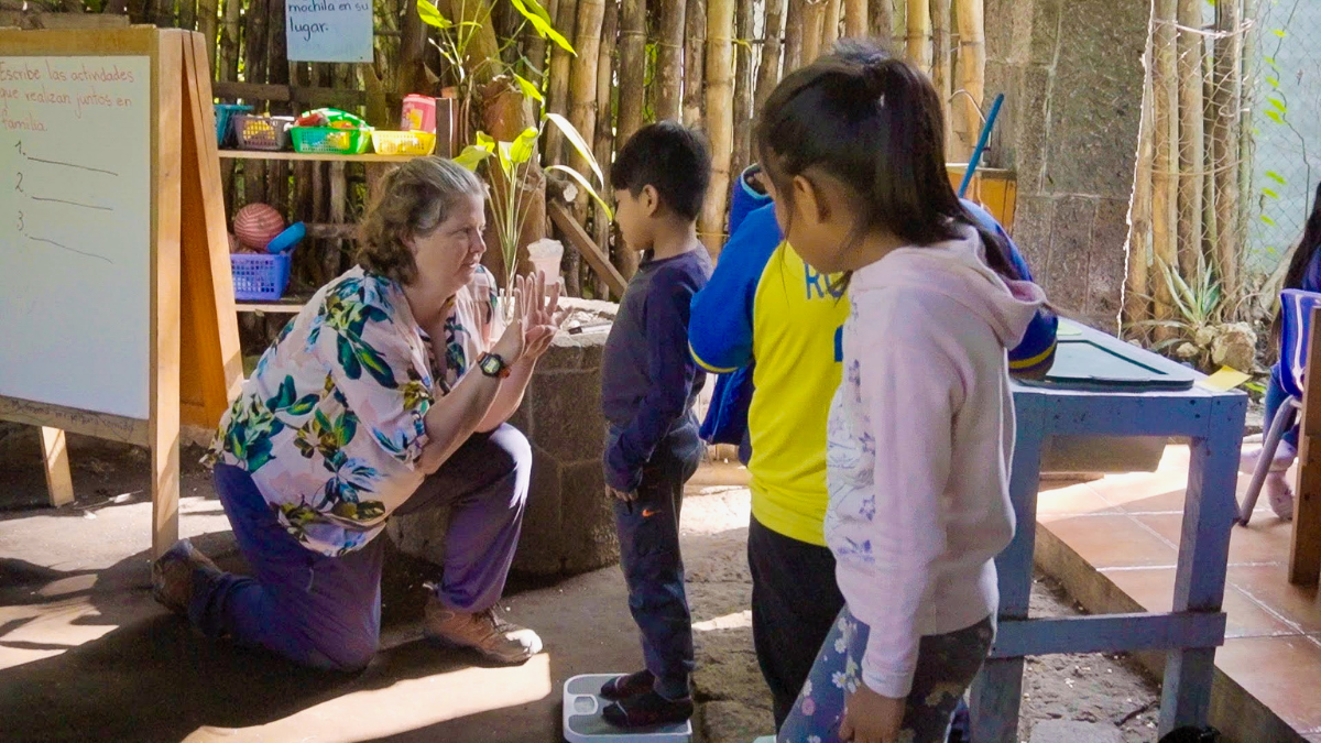 Woman crouching and engaging warmly with young children in an outdoor classroom, with one child standing on a scale and others waiting in line, surrounded by bamboo walls, a whiteboard with Spanish writing, and colorful educational supplies