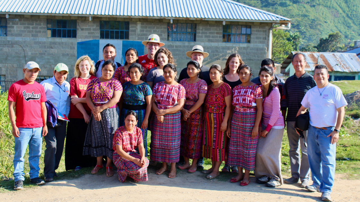 Large group of teachers and community members pose outdoors in front of a rural school many women wearing colorful Mayan huipiles on a sunny day
