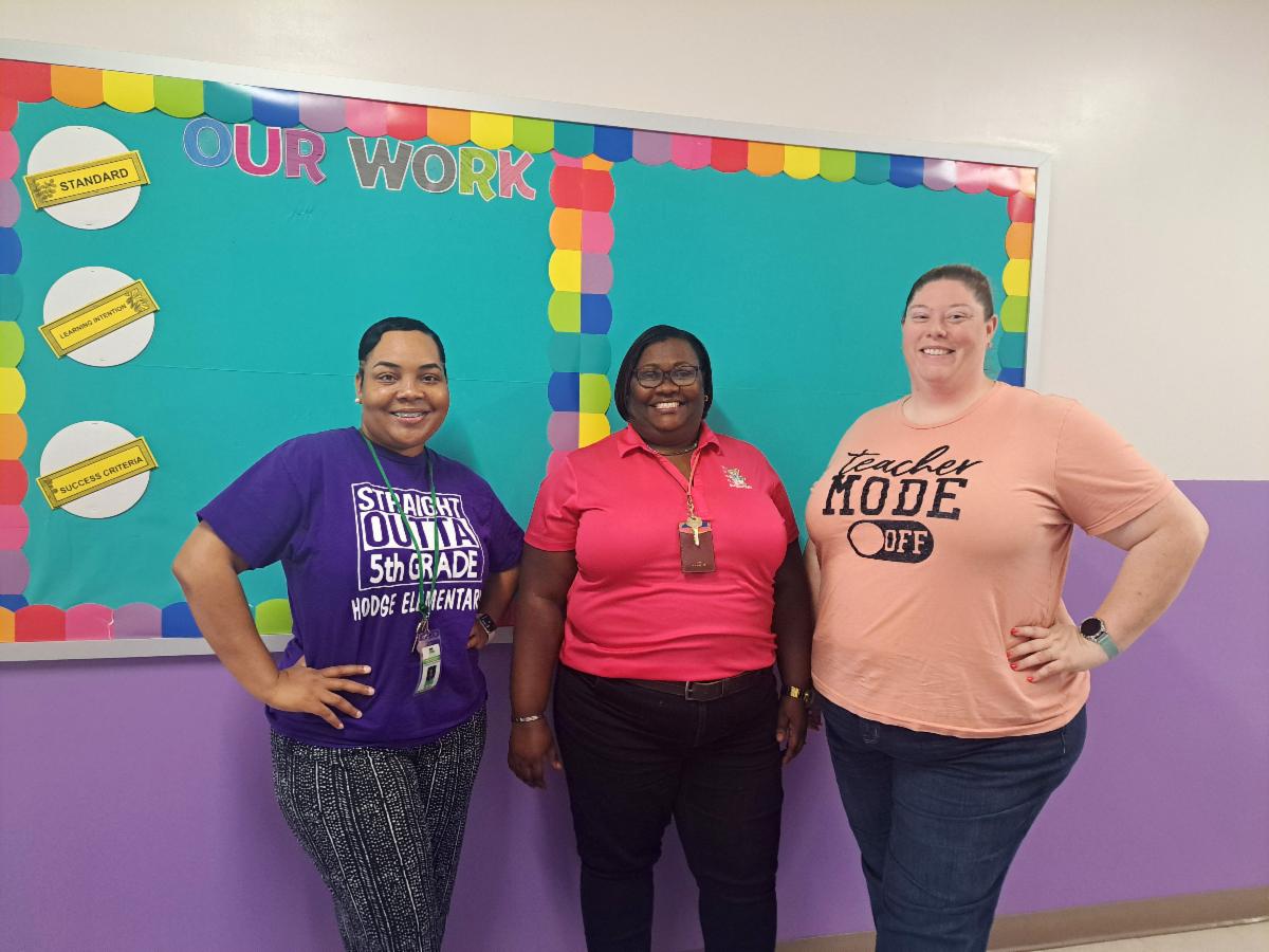 Three women pose in front of a school bulletin board