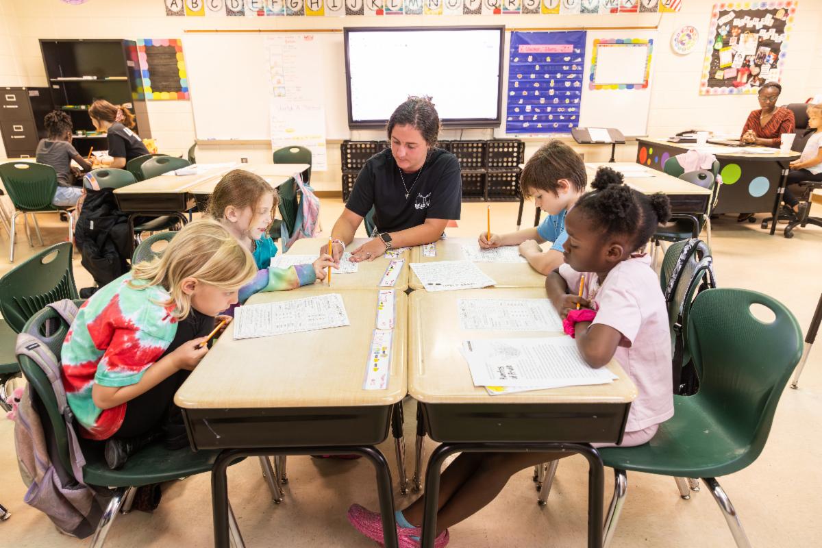 photo of four children with papers at a cluster of desks with a young woman
