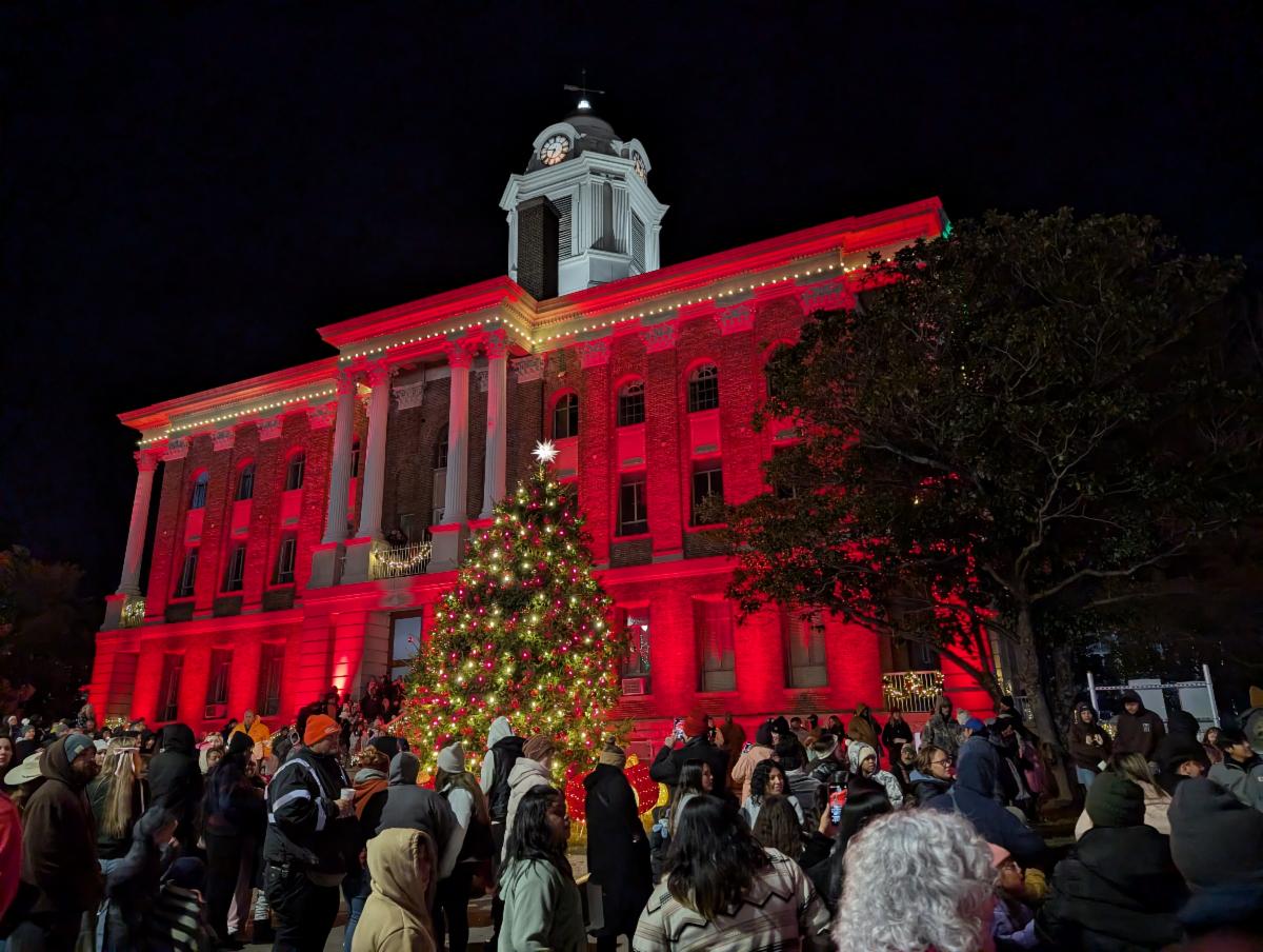 Bedford County Courthouse, flooded in red lighting with white Christmas lights as trim, and in the foreground the decorated and lit Christmas tree, with a crowd of spectators.