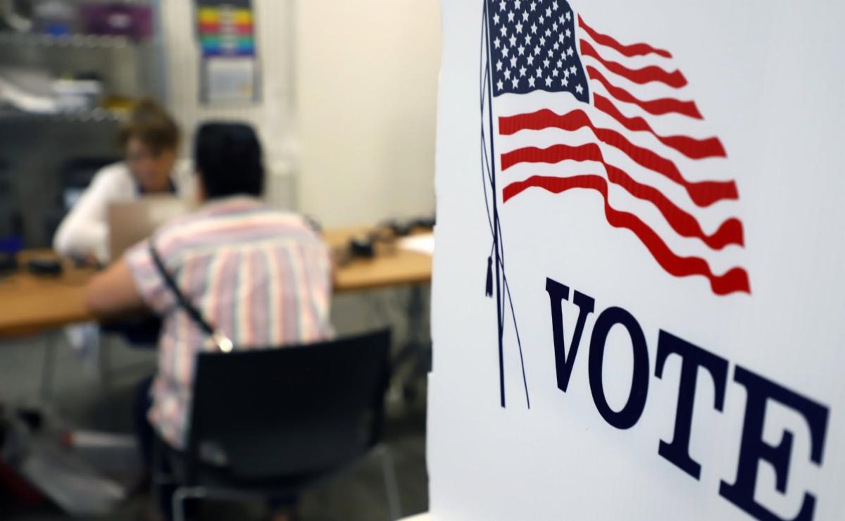 A voter sits across from an election worker at a table, with an American flag and VOTE sign in the foreground.