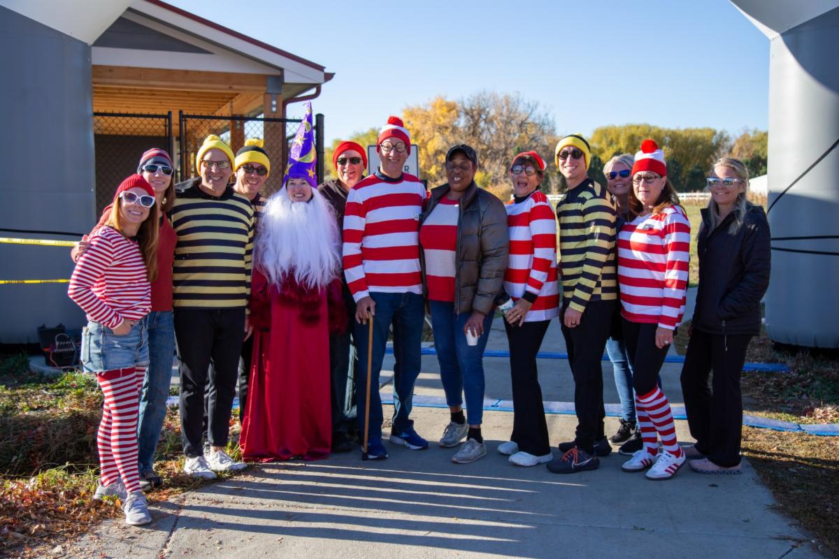 Eleven people pose together outdoors wearing colorful striped costumes, hats and accessories.