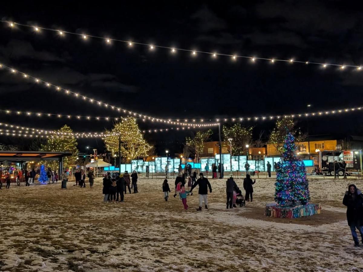 Visitors walk through a snow-covered plaza beneath strings of lights and illuminated trees at a winter celebration.
