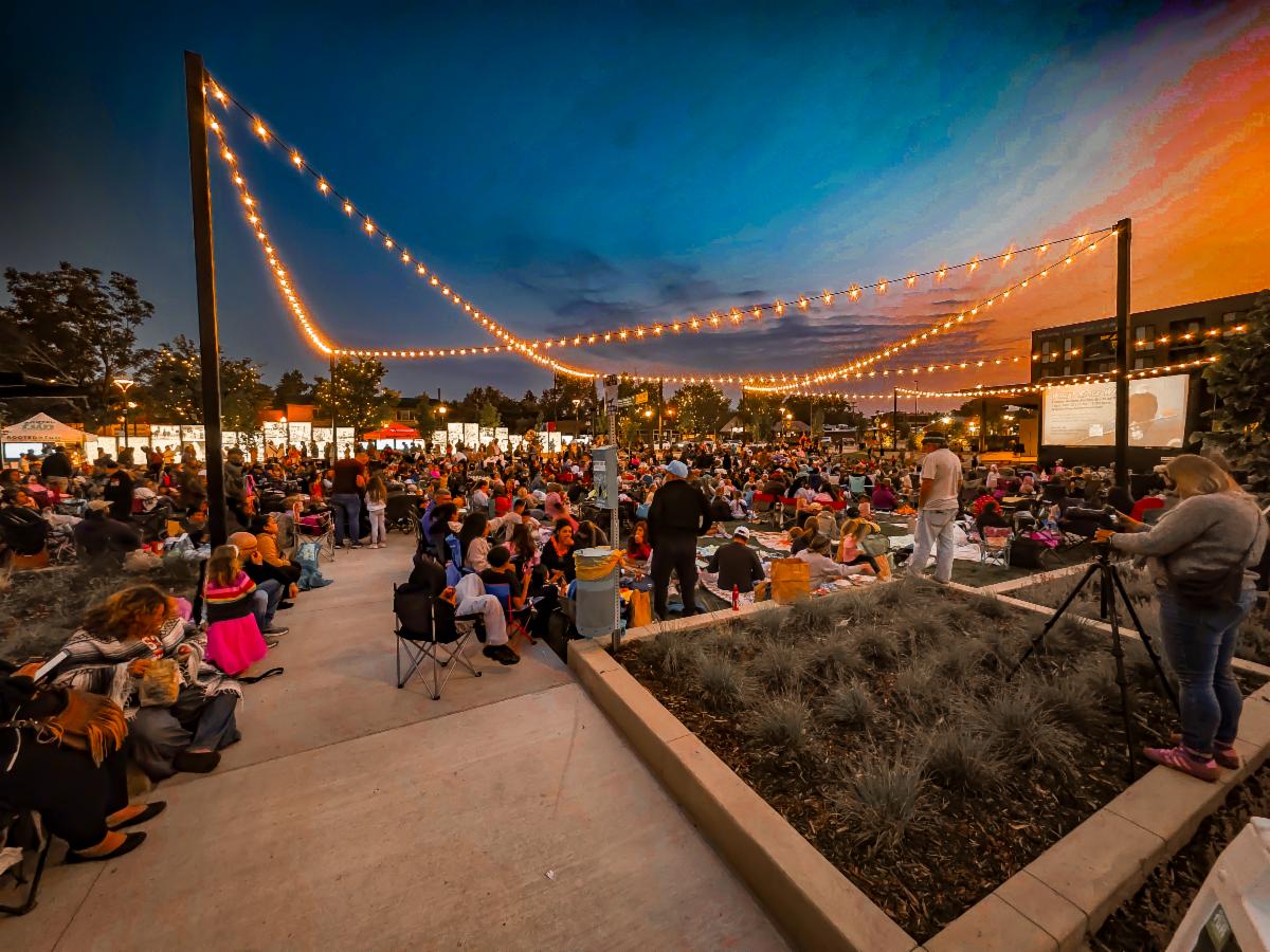 Outdoor movie night at dusk with people seated under string lights near a projection screen.