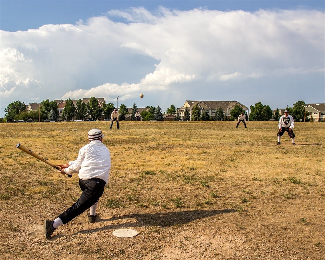 Vintage baseball players compete on an open field, with a batter swinging at the ball.