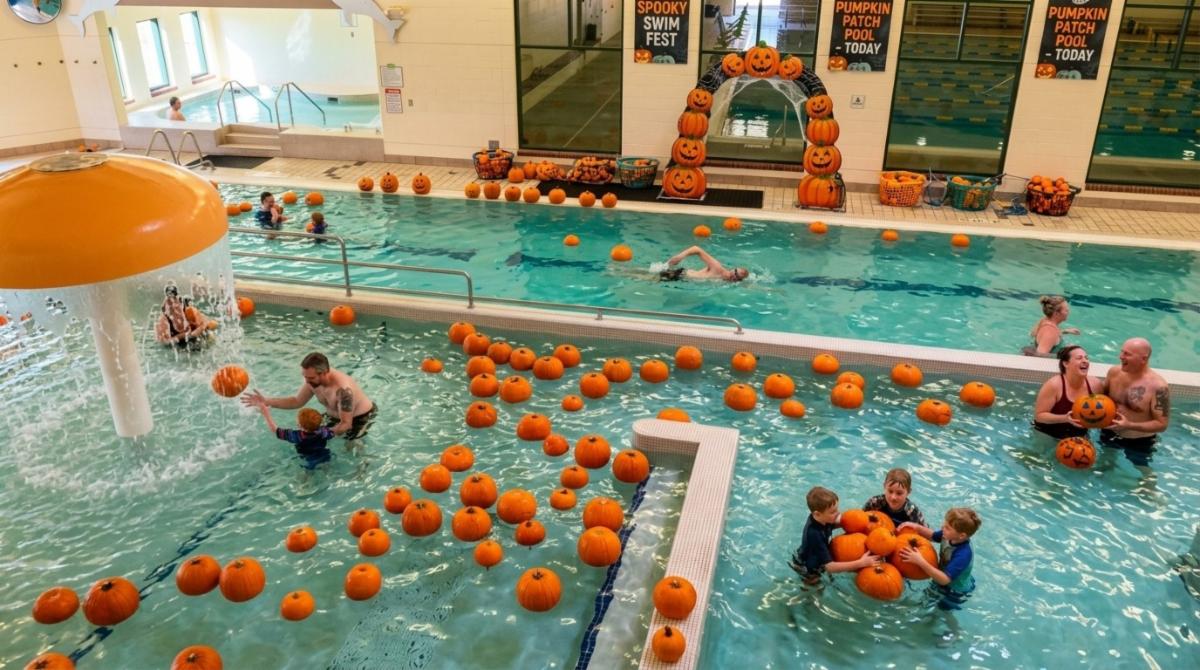 Families swim among floating pumpkins during an indoor pumpkin patch pool event.