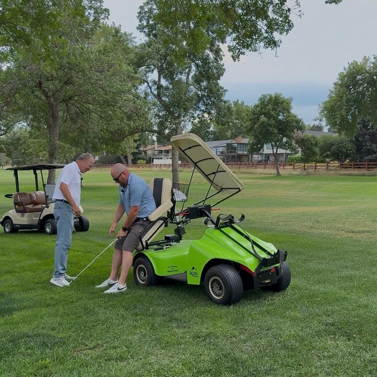 Two men on a golf course beside a bright green adaptive golf cart; one man is seated in the cart while holding a golf club and the other stands nearby.