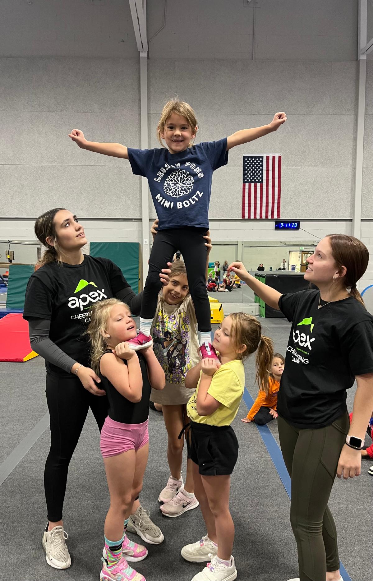 Children and cheer coaches pose in a gym, with one child lifted high in a cheer stunt.