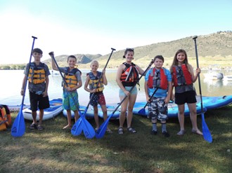 Children standing with paddles beside kayaks by a lake.