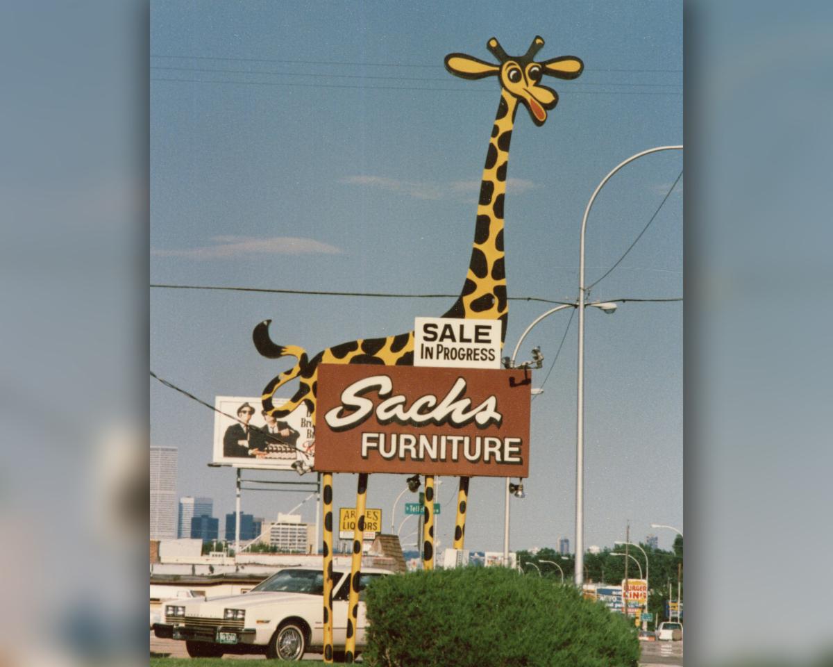 Vintage street photo of a large giraffe-shaped Sachs Furniture sign with a ‘Sale in Progress’ placard, cars, power lines, and a city skyline in the background.