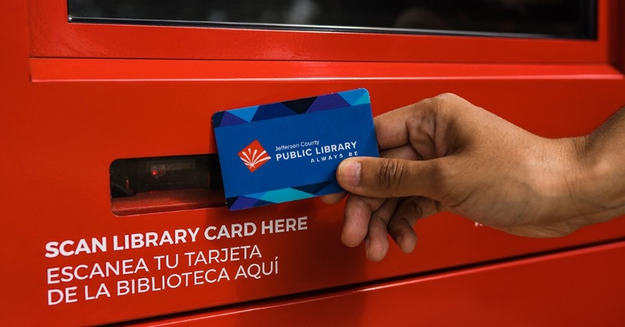 A hand scans a Jefferson County Public Library card at a red self-service kiosk.