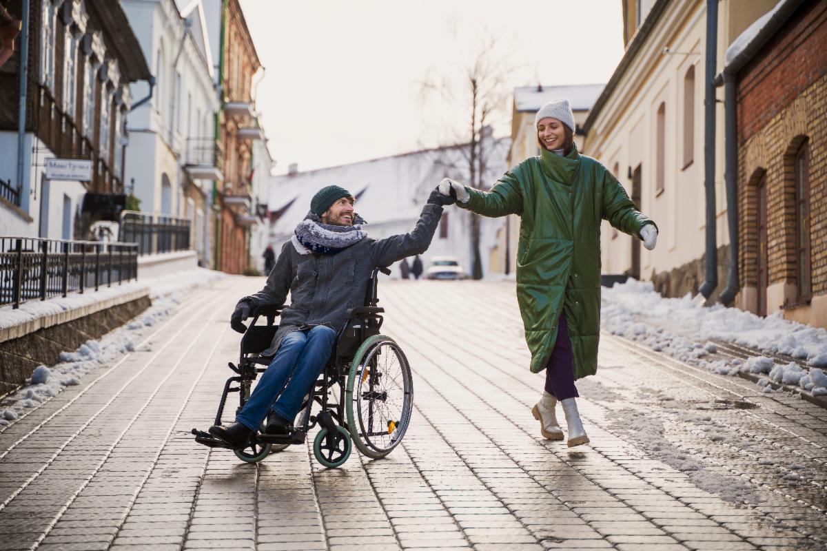 Photo of man in a wheelchair holding the hand of a woman as they walk down the street. They are in an urban area on a stone pathway. They are wearing winter coats and hats.