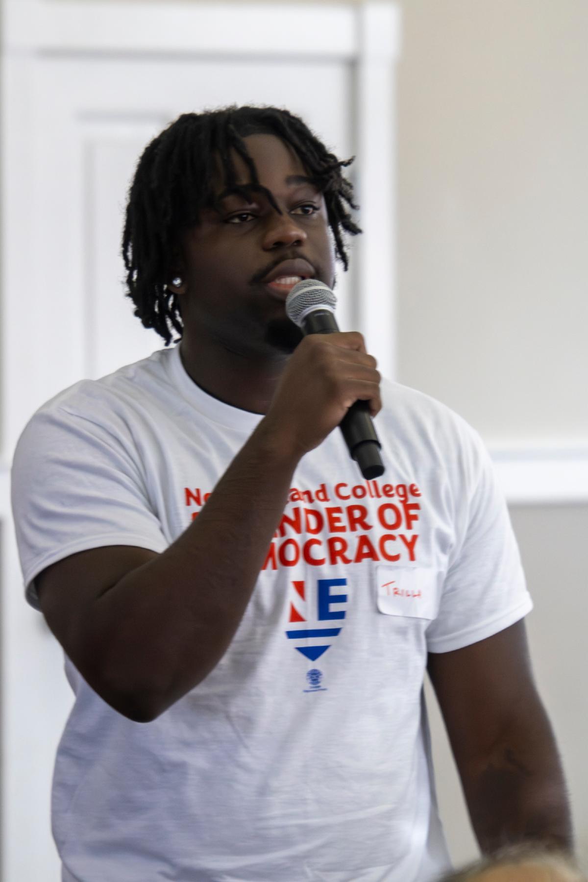 A young black man speaks at a mic. He's wearing a shirt that says "defenders of democracy" in big block letters.