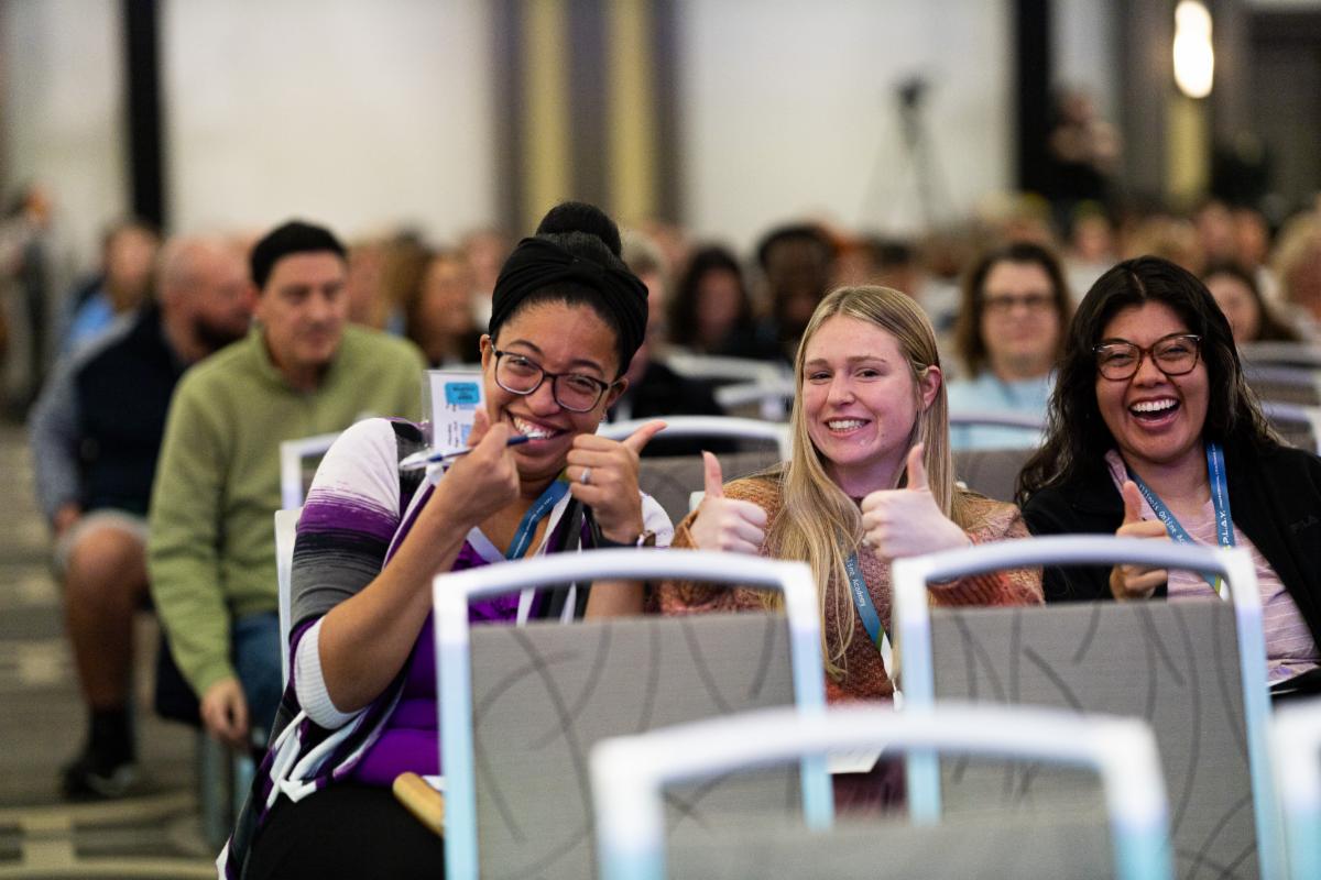 Three professionals smiling with their thumbs up while sitting in a crowded conference room.