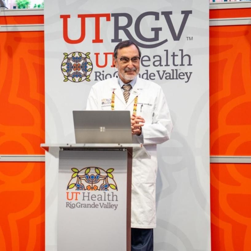 A person in a white lab coat stands at a podium with a laptop, smiling, against a backdrop with "UTRGV UT Health Rio Grande Valley" branding.
