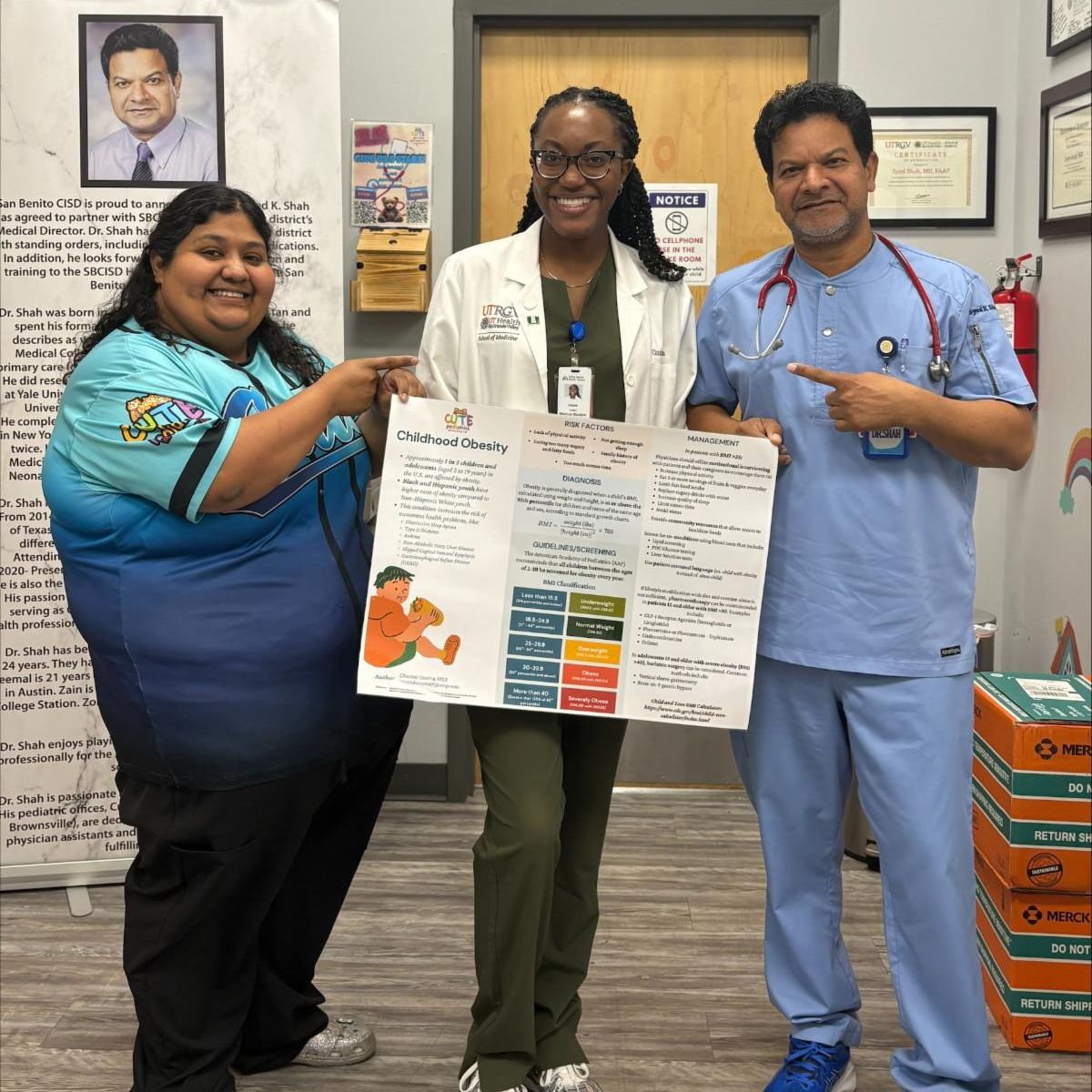 Three people smile while holding a "Childhood Obesity" poster in a pediatric clinic. The wall has colorful decorations and a poster of bears.