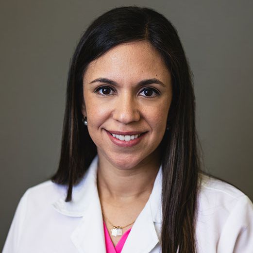 Headshot of a smiling lady wearing a white medical white coat. She is wearing a bright pink shirt under, with a silver necklace. 