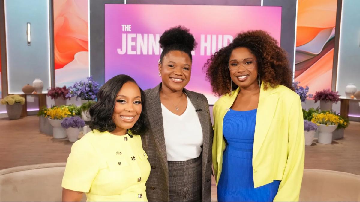 Three women stand smiling on a colorful talk show set. Two wear yellow outfits, and one wears a plaid suit. The atmosphere is warm and inviting.