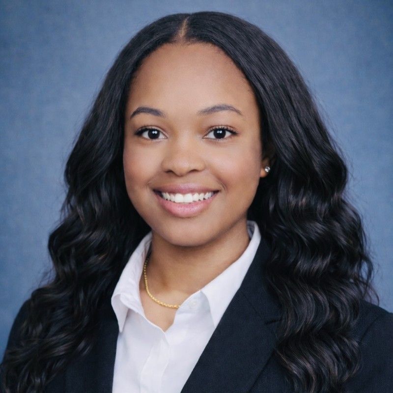Smiling woman with long, wavy hair wearing a black blazer, white shirt, and gold necklace against a blue background, conveying professionalism and warmth.