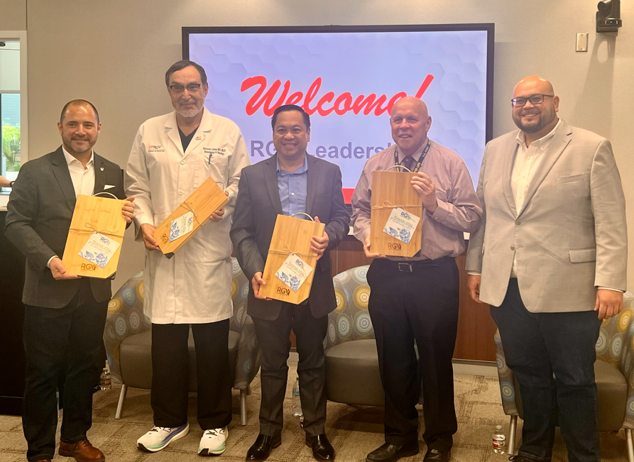 Five men smiling in a conference room holding wooden cutting boards. A screen in the background displays "Welcome! RGV Leadership." The mood is celebratory.