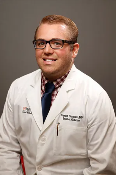 A smiling man in glasses wearing a white coat over a red checkered shirt and blue tie. The coat has medical embroidery. Neutral, professional tone.