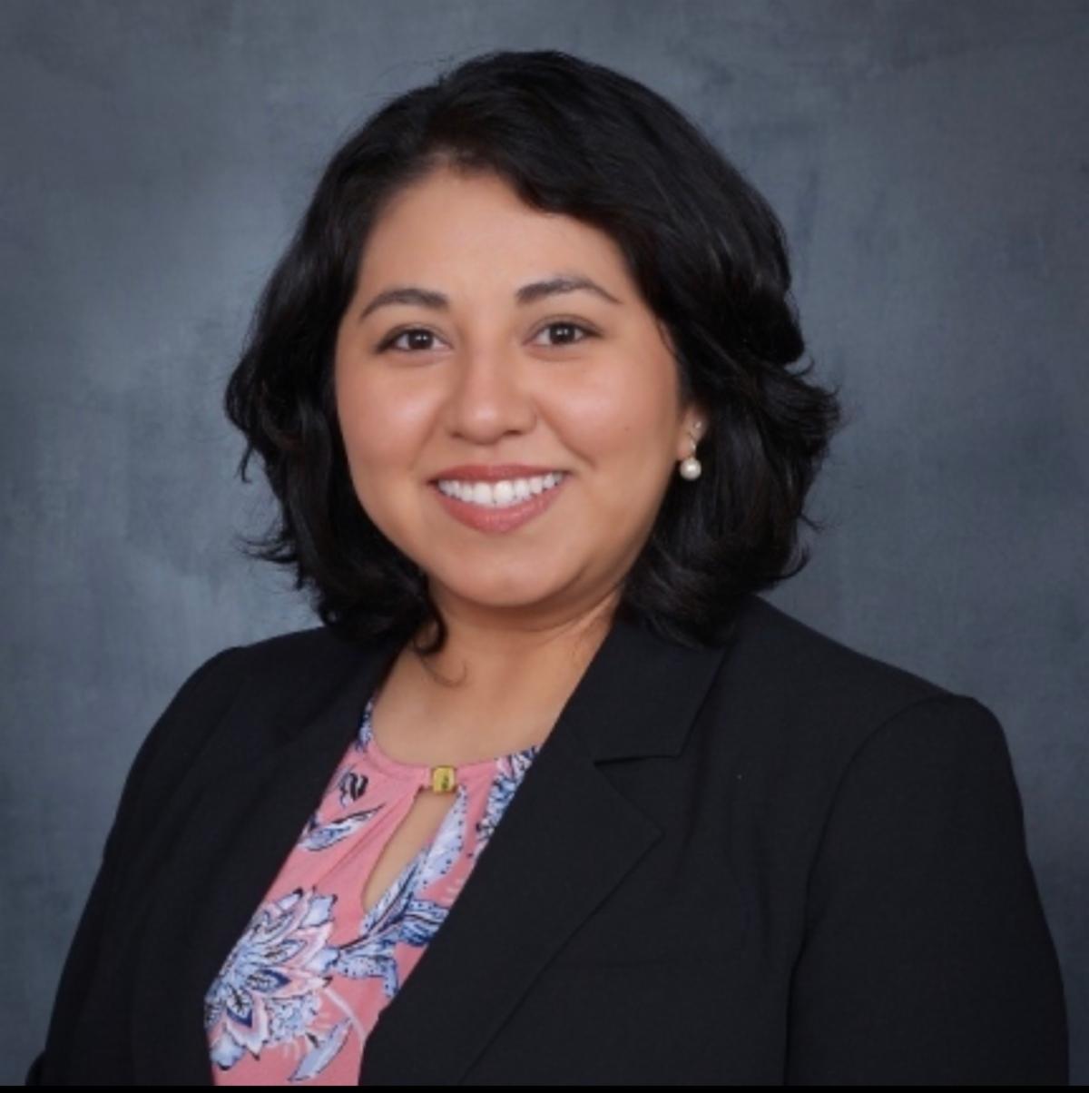 Smiling woman in a black blazer and pink floral blouse against a gray background. She has shoulder-length dark hair and is wearing pearl earrings.